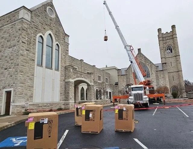 A crane lifts a box toward a stone church. Several boxes sit in a parking lot. Overcast sky.