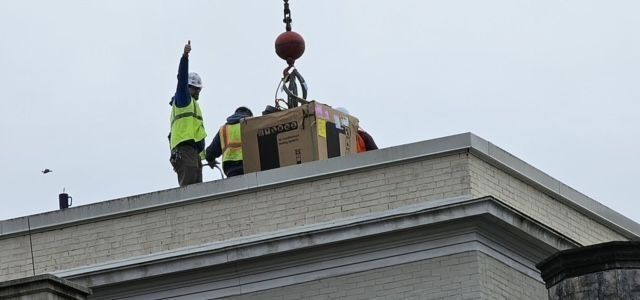 Workers on a rooftop lift a large box with a crane on an overcast day. One worker signals.