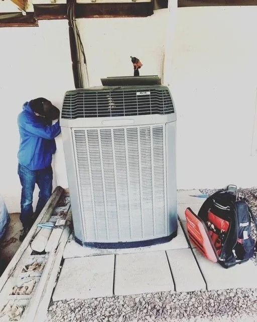 Man in blue jacket beside an AC unit, leaning with head down. Toolbox and materials in view.