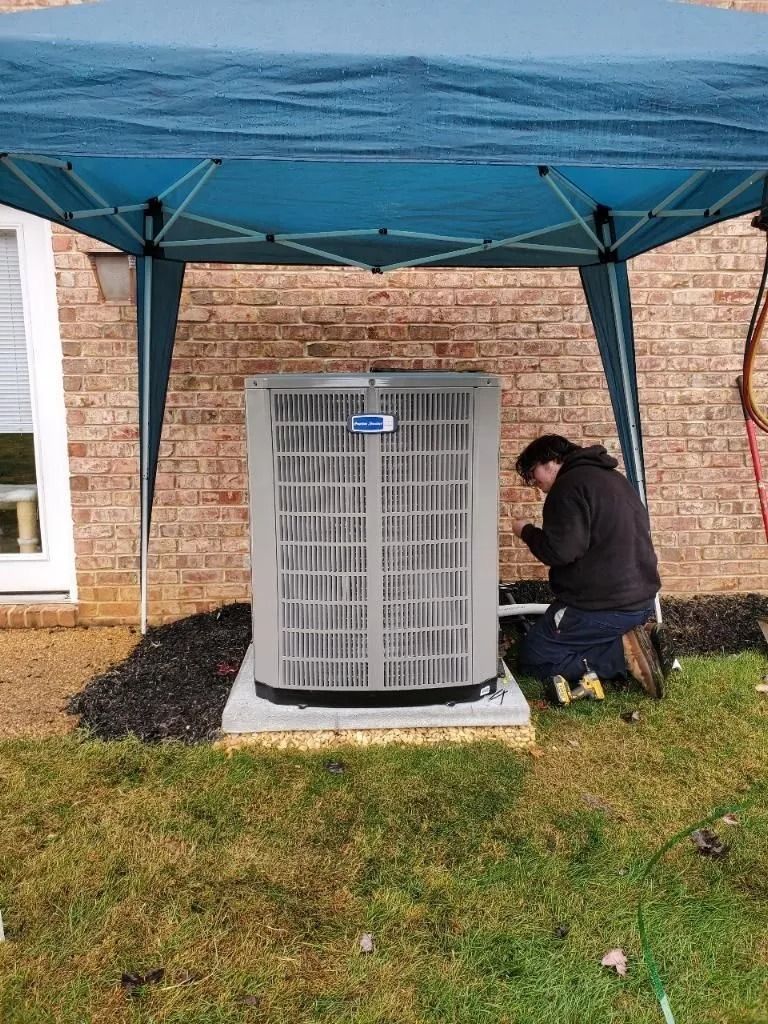 Man working on an AC unit under a blue canopy next to a brick building, set on grass.