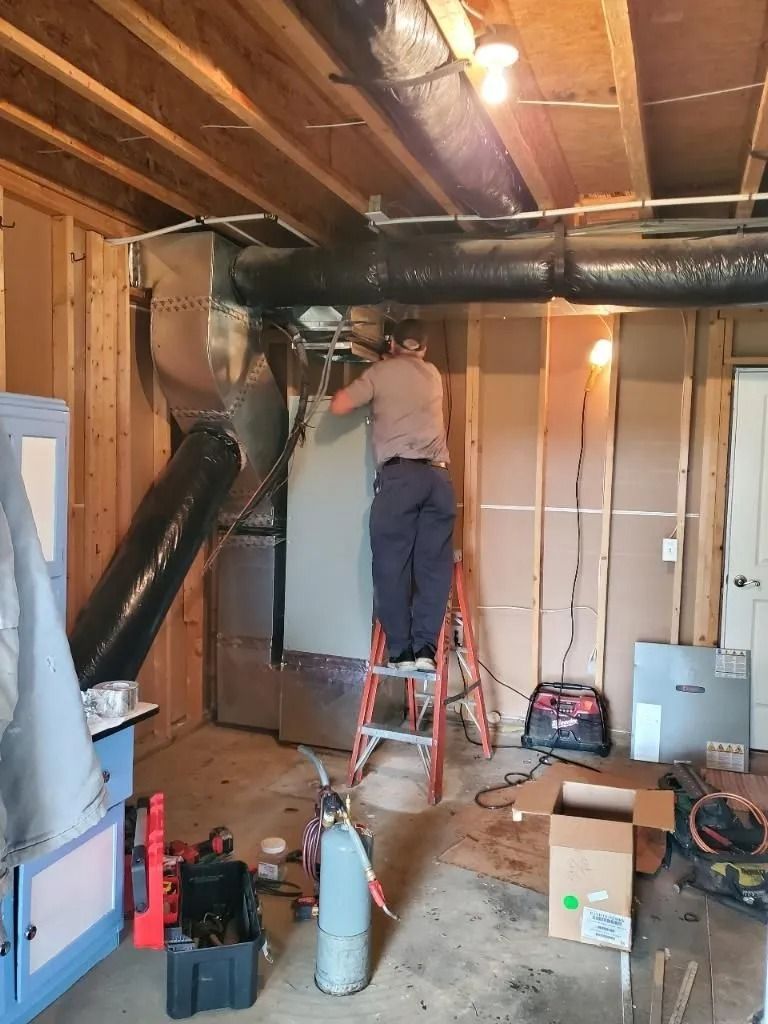 A person on a ladder installs ductwork in a basement. Exposed studs, tools, and HVAC equipment are visible.