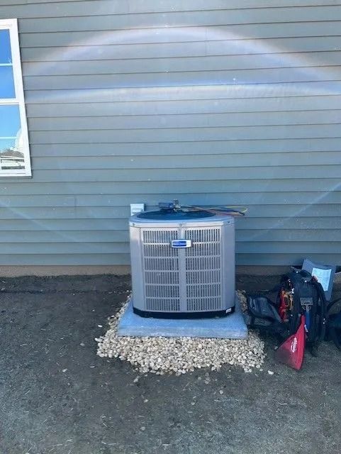 An air conditioning unit on a concrete pad, surrounded by gravel, next to a blue-sided house.