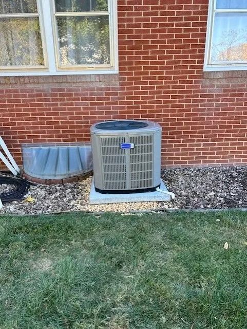 An air conditioning unit outside a brick building, beside a window well.