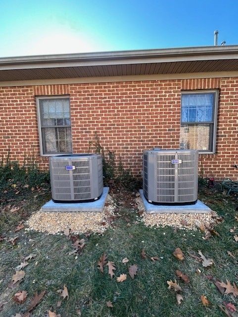 Two air conditioning units on concrete pads next to a brick building and windows, surrounded by gravel and grass.