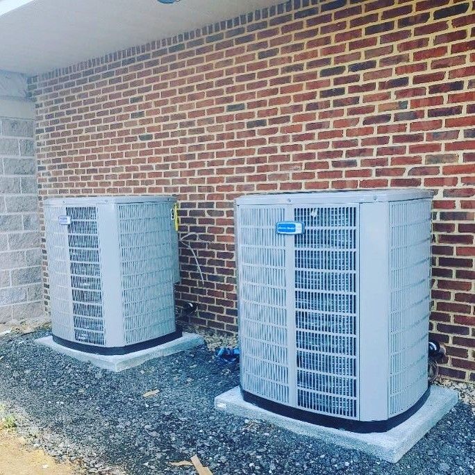 Two grey air conditioning units sit on concrete pads against a brick wall.