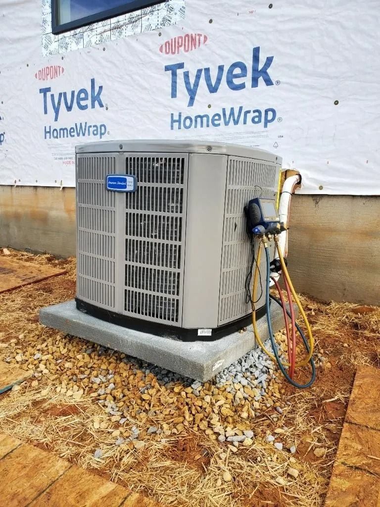 Air conditioning unit on a concrete pad, being serviced with hoses. Outside, near a building under construction.