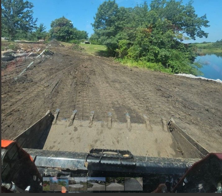 View from the cab of a skid steer tractor moving dirt; muddy ground, trees, and water in the background.
