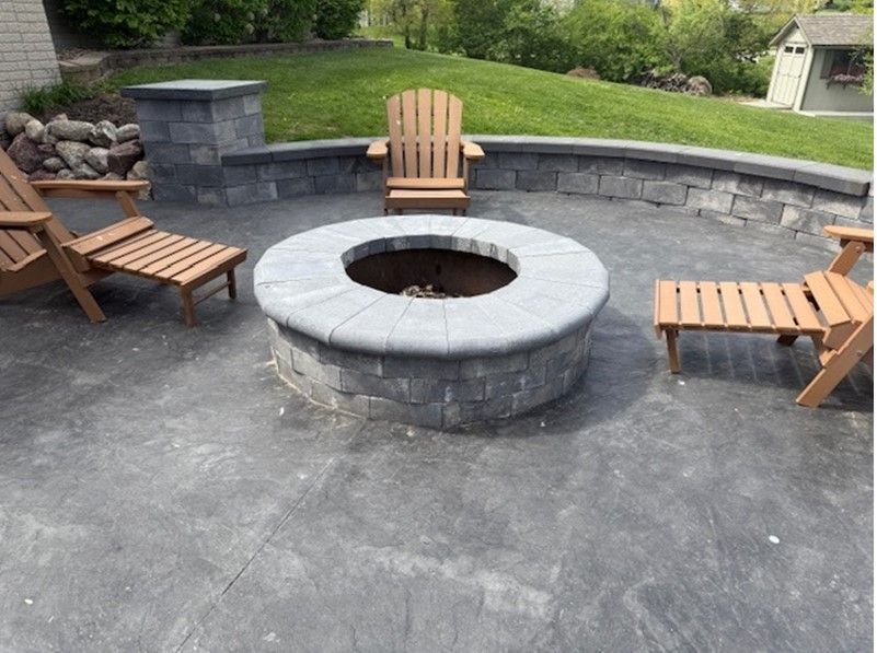 Stone fire pit on a patio with three wooden chairs; green grass in the background.