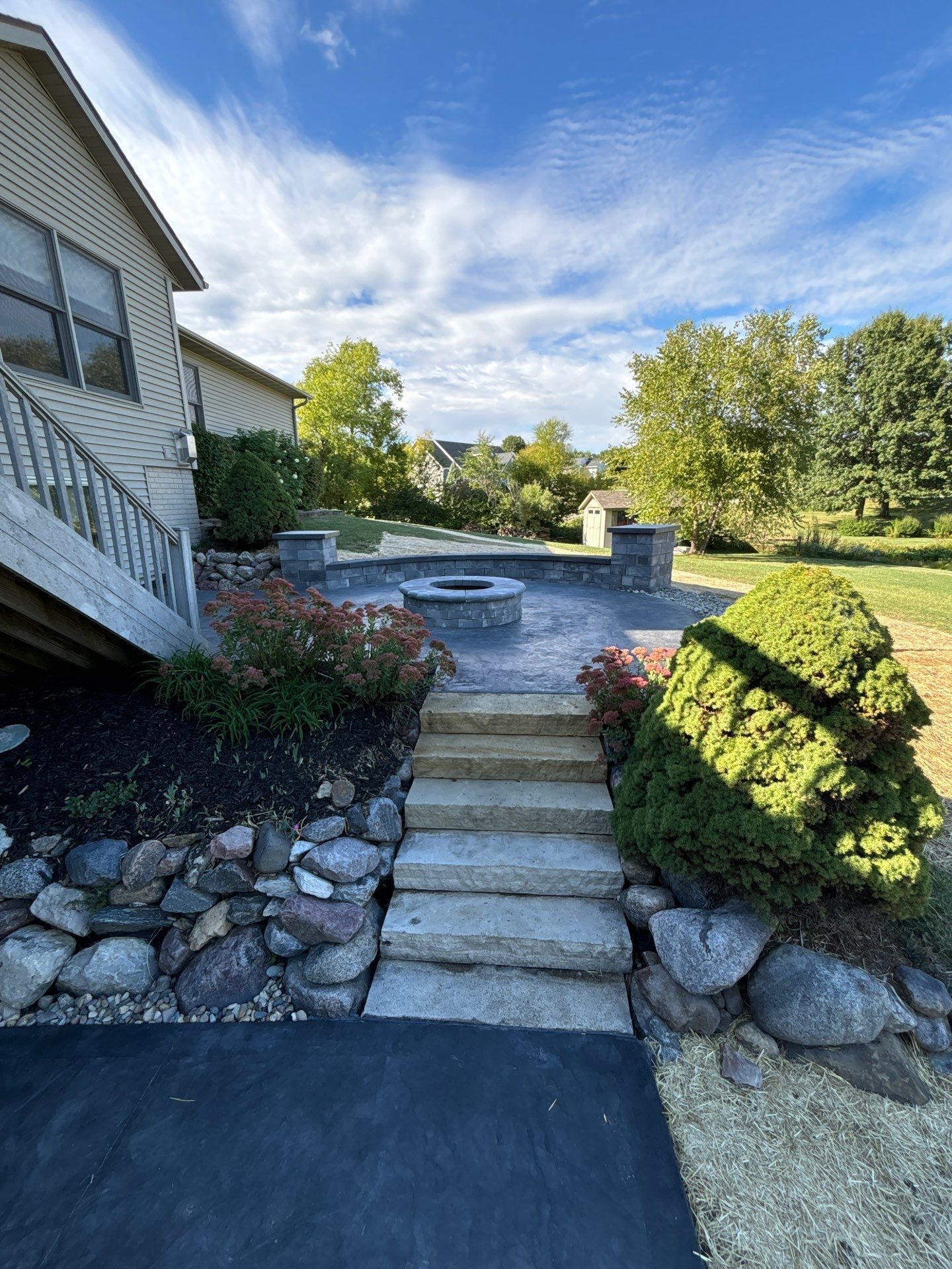 Stone patio with fire pit, steps, and landscaping next to a house under a blue sky.