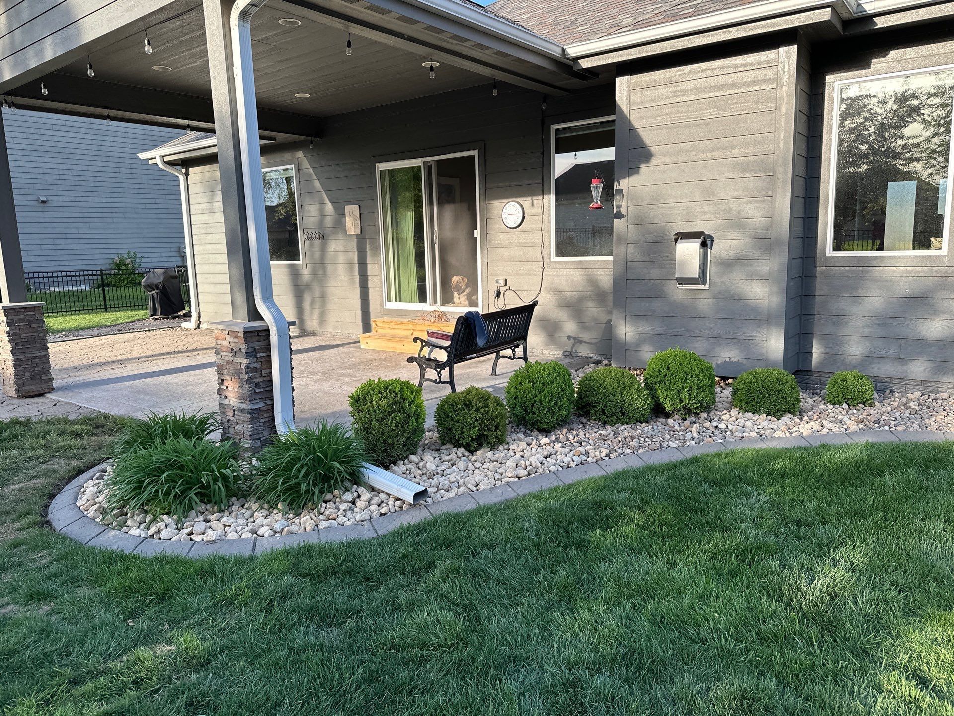 Backyard patio with green bushes, rocks, and a bench along a house's exterior.