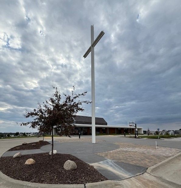 A tall white cross stands in front of a modern building under a cloudy sky.