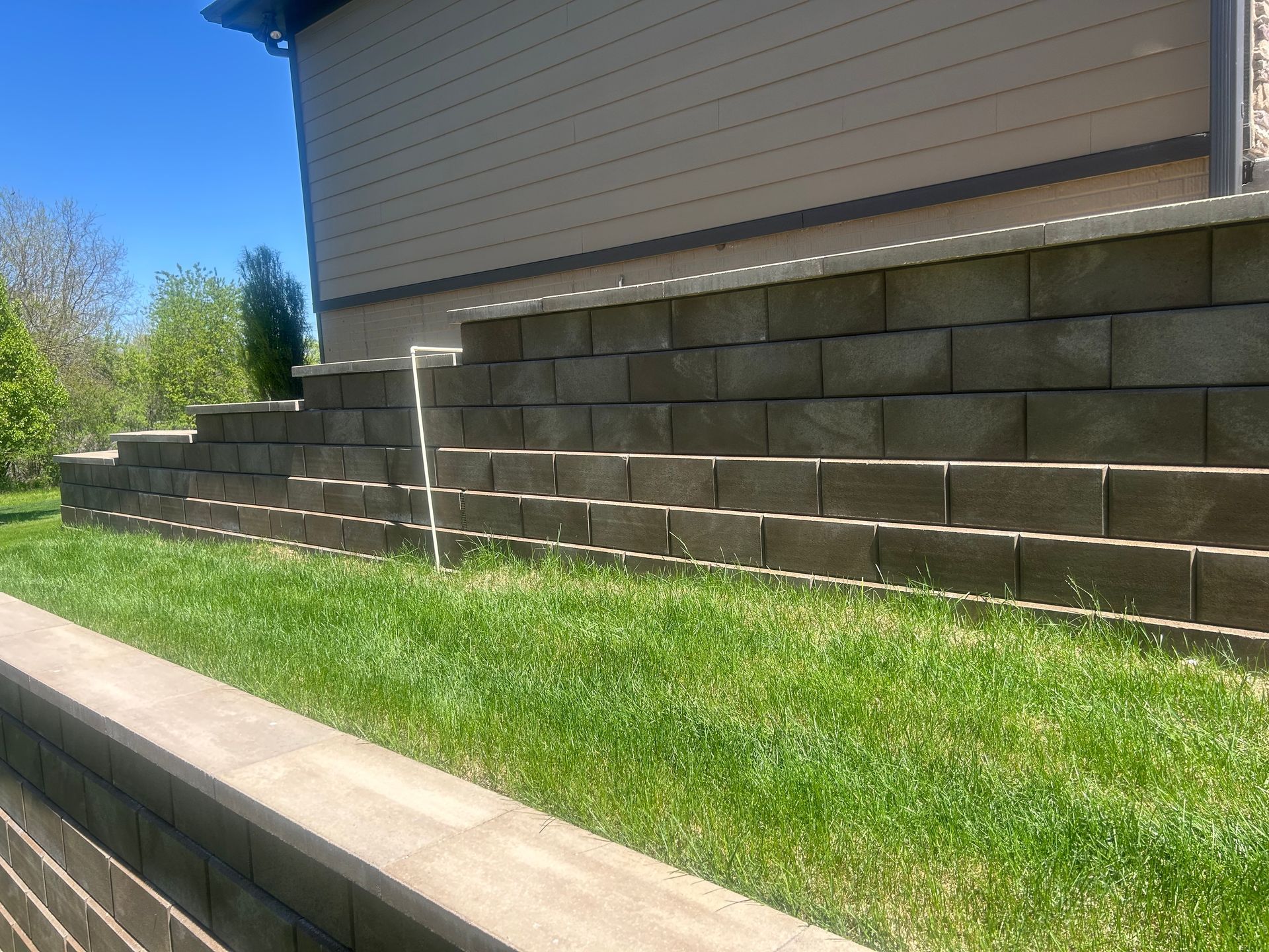 Stone retaining wall with a stepped design, green grass, and a beige house under a blue sky.