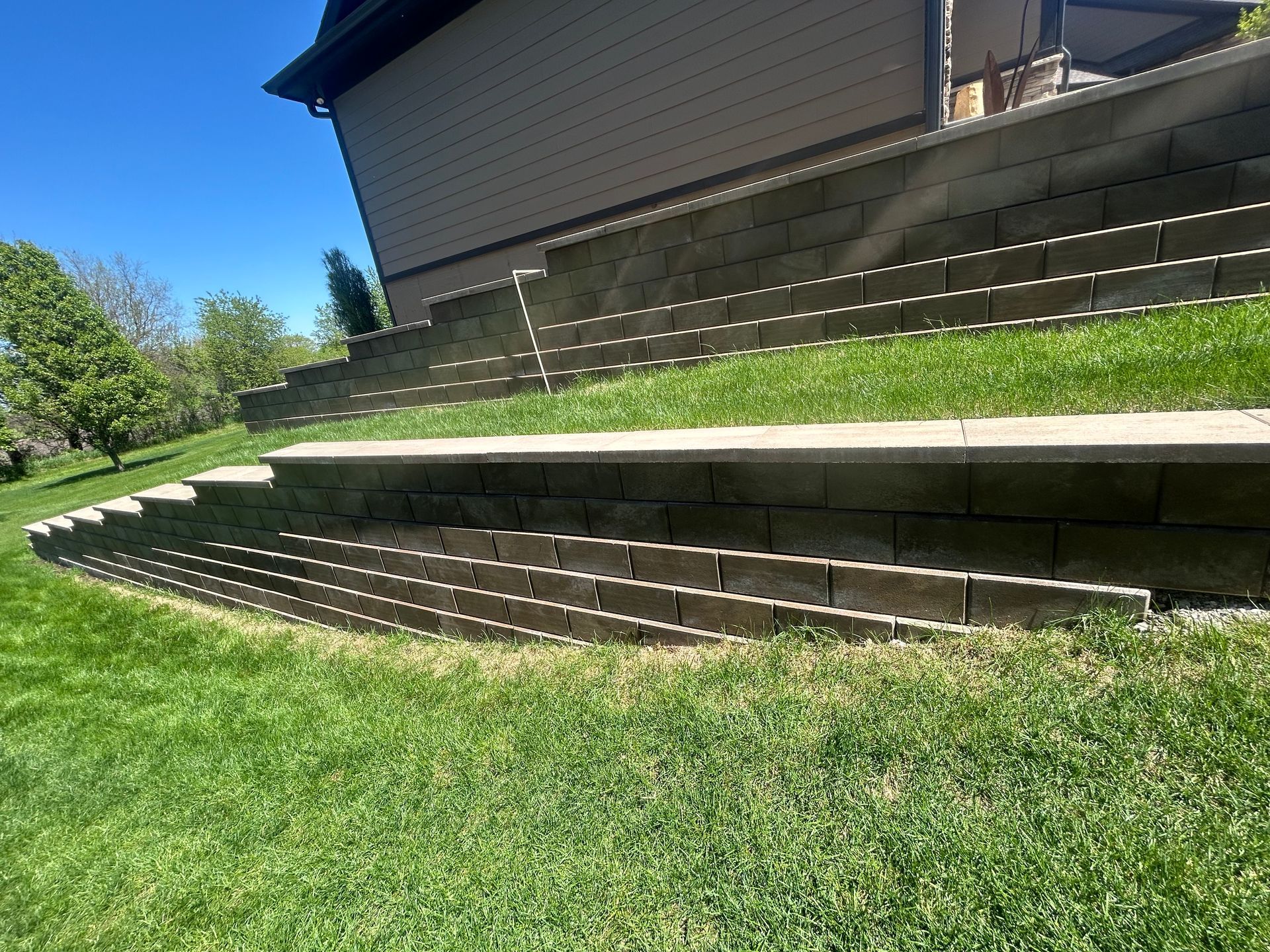Stone retaining wall with steps, grass lawn, and house exterior on a sunny day.