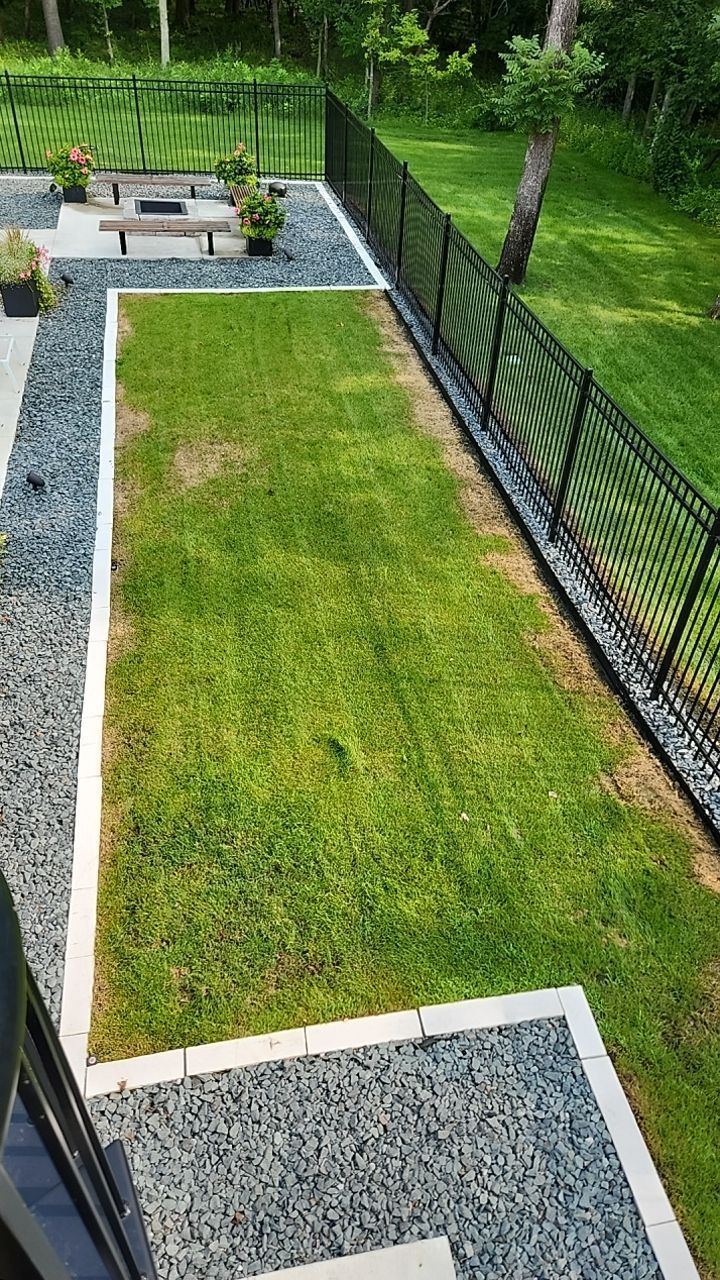 Overhead view of backyard: green grass area bordered by dark gravel, concrete, and black fence.