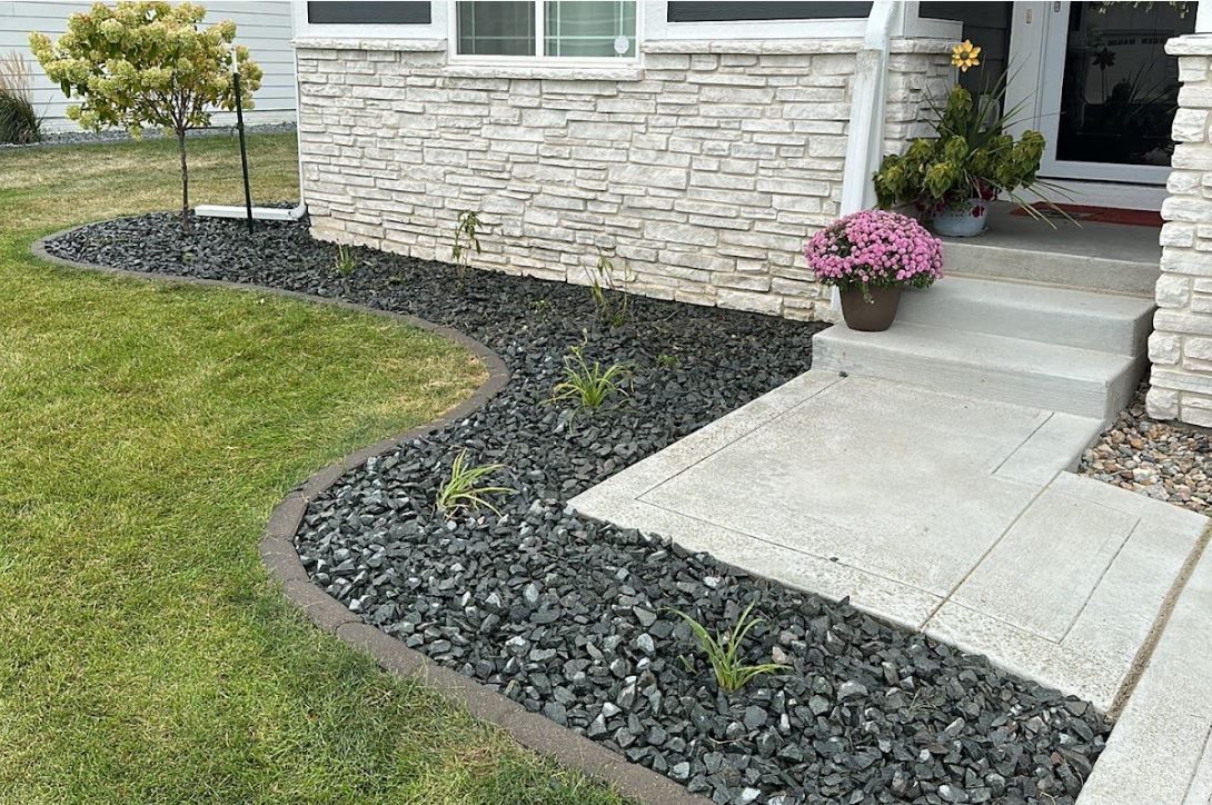 Front yard landscaping with black stones, small plants, and a concrete walkway.