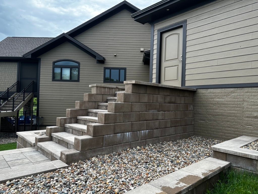 Stone steps leading up to a beige building with a dark roof. Gravel and pavers are in front of the steps.