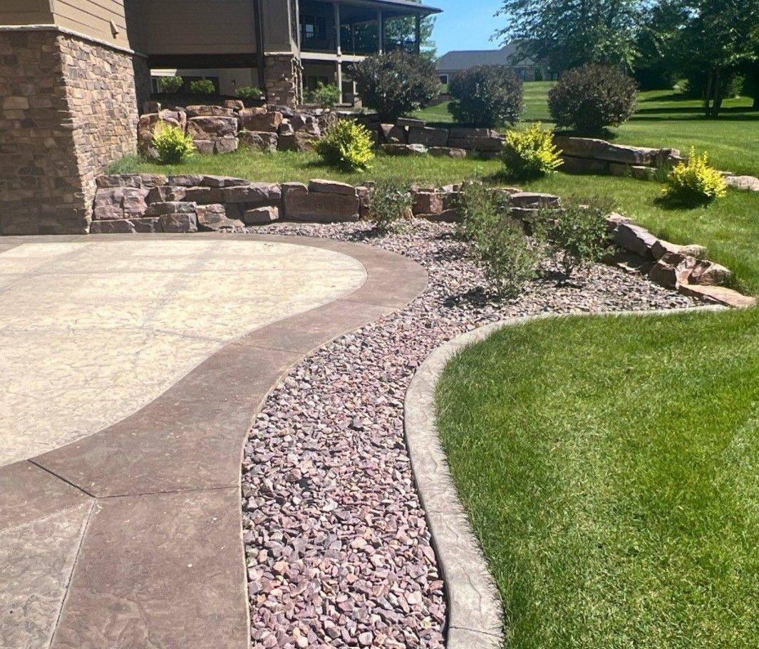 Curving concrete path with red gravel bed next to green lawn, leading towards a stone retaining wall and building.