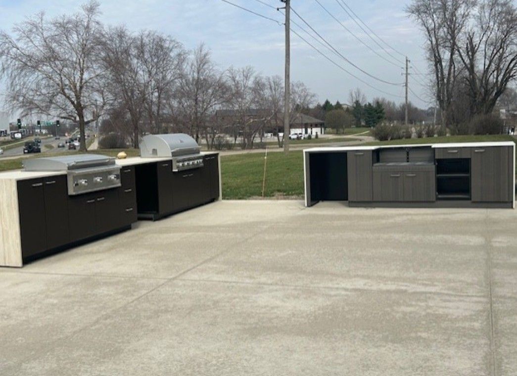 Two dark gray, modern outdoor kitchen islands with stainless steel grills sit on a concrete patio outdoors.