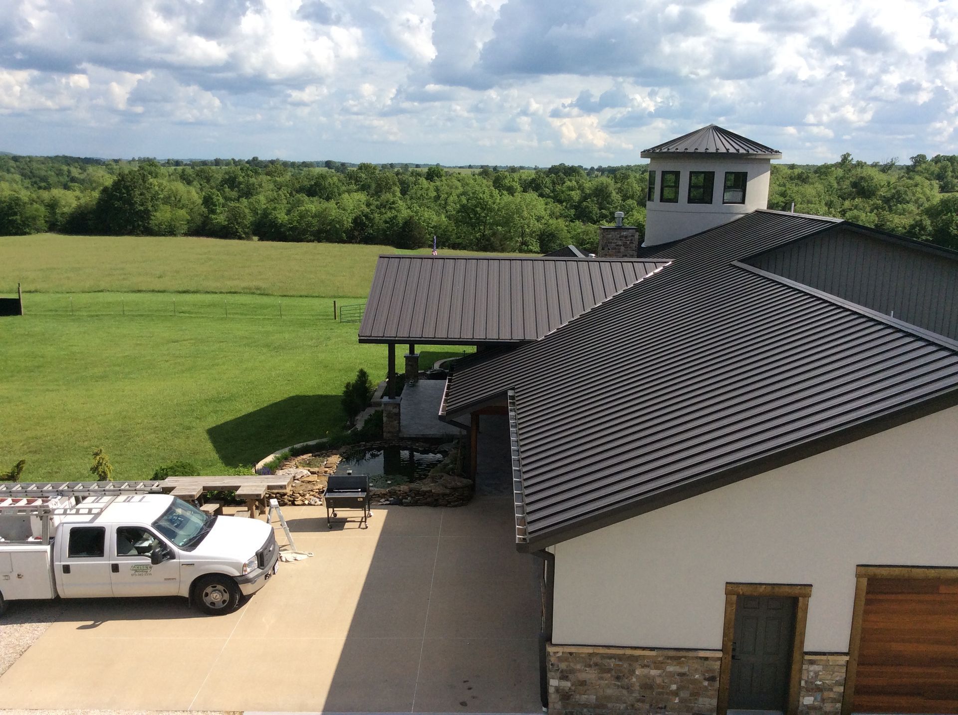 White pickup truck by a house with a dark roof, overlooking green fields and trees under a blue sky
