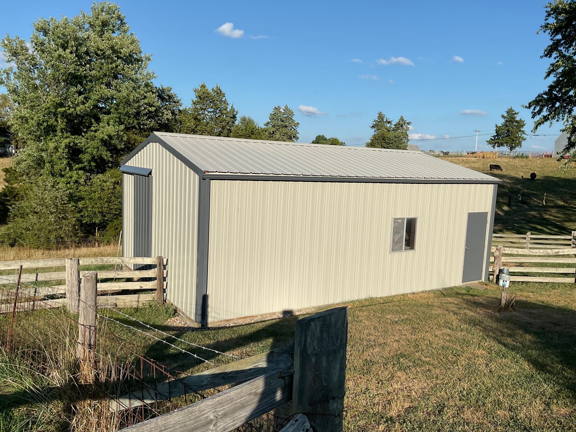 Small beige metal shed with a gray roof in a grassy rural yard under a blue sky.