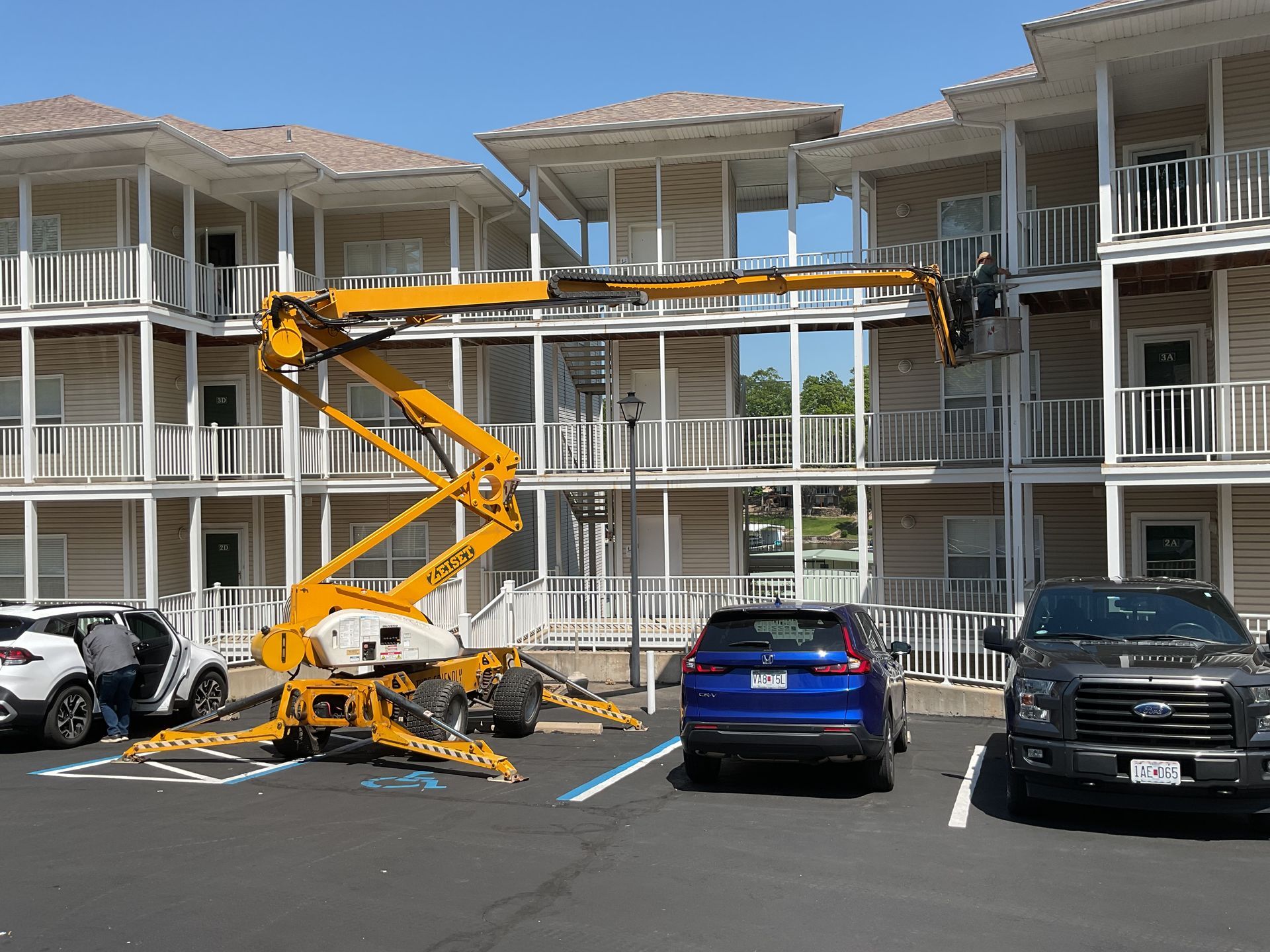 Yellow lift parked in front of a white apartment building, with cars in the lot.