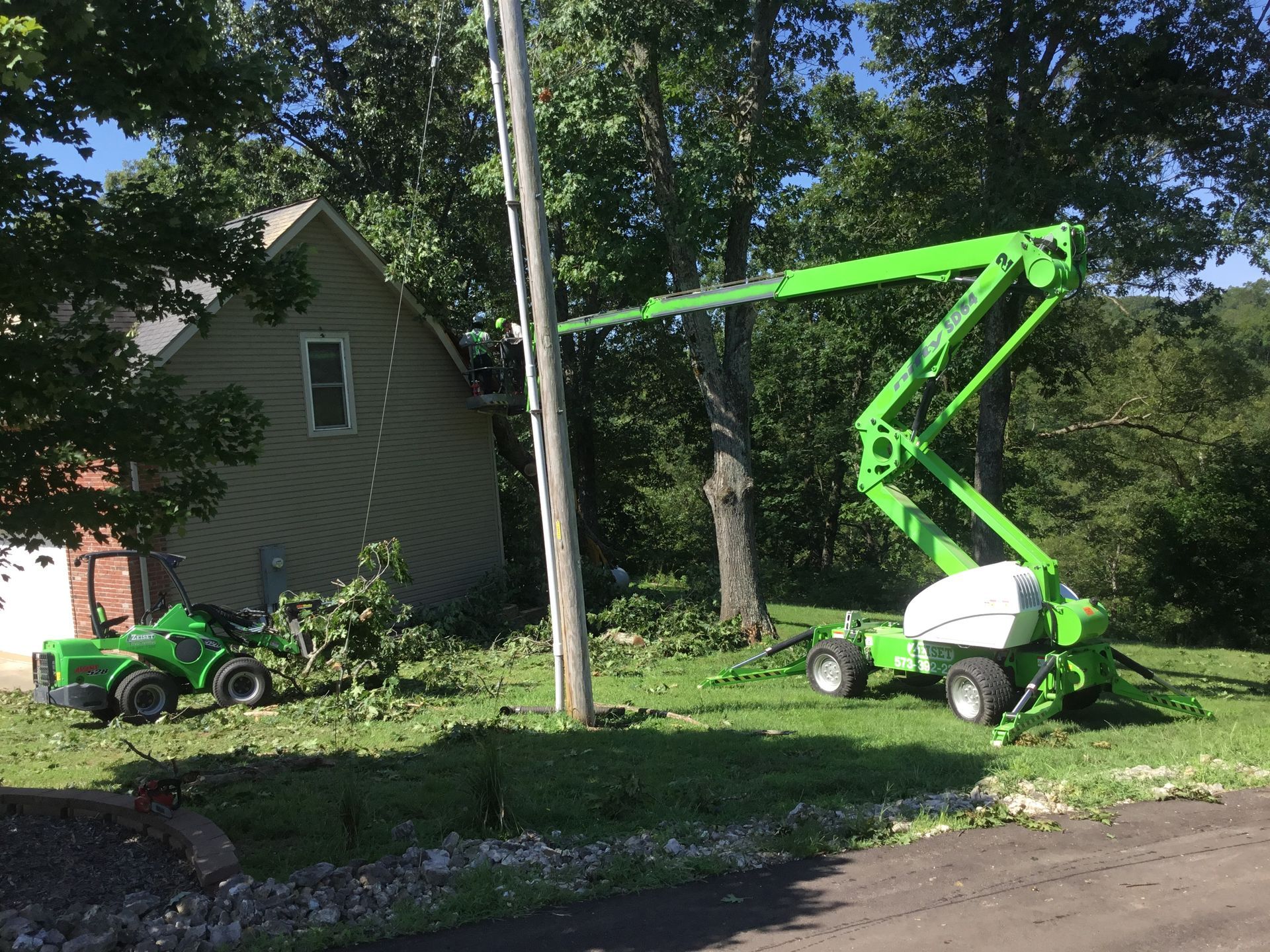 Green utility lifts near a house, one by a pole and one on grass, with trees and a partly visible truck.