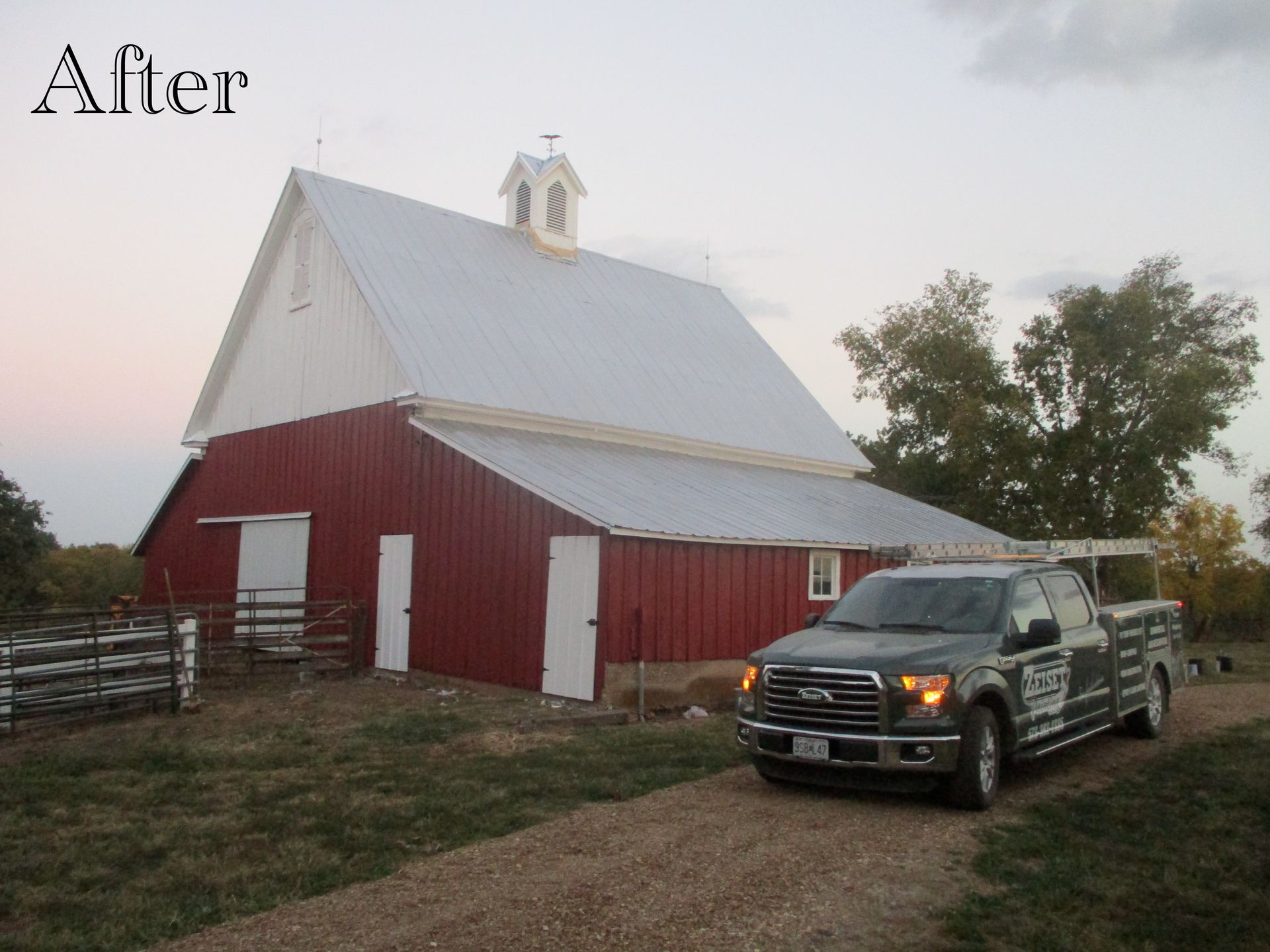 Red barn with white roof and a black pickup truck parked beside it at dusk.