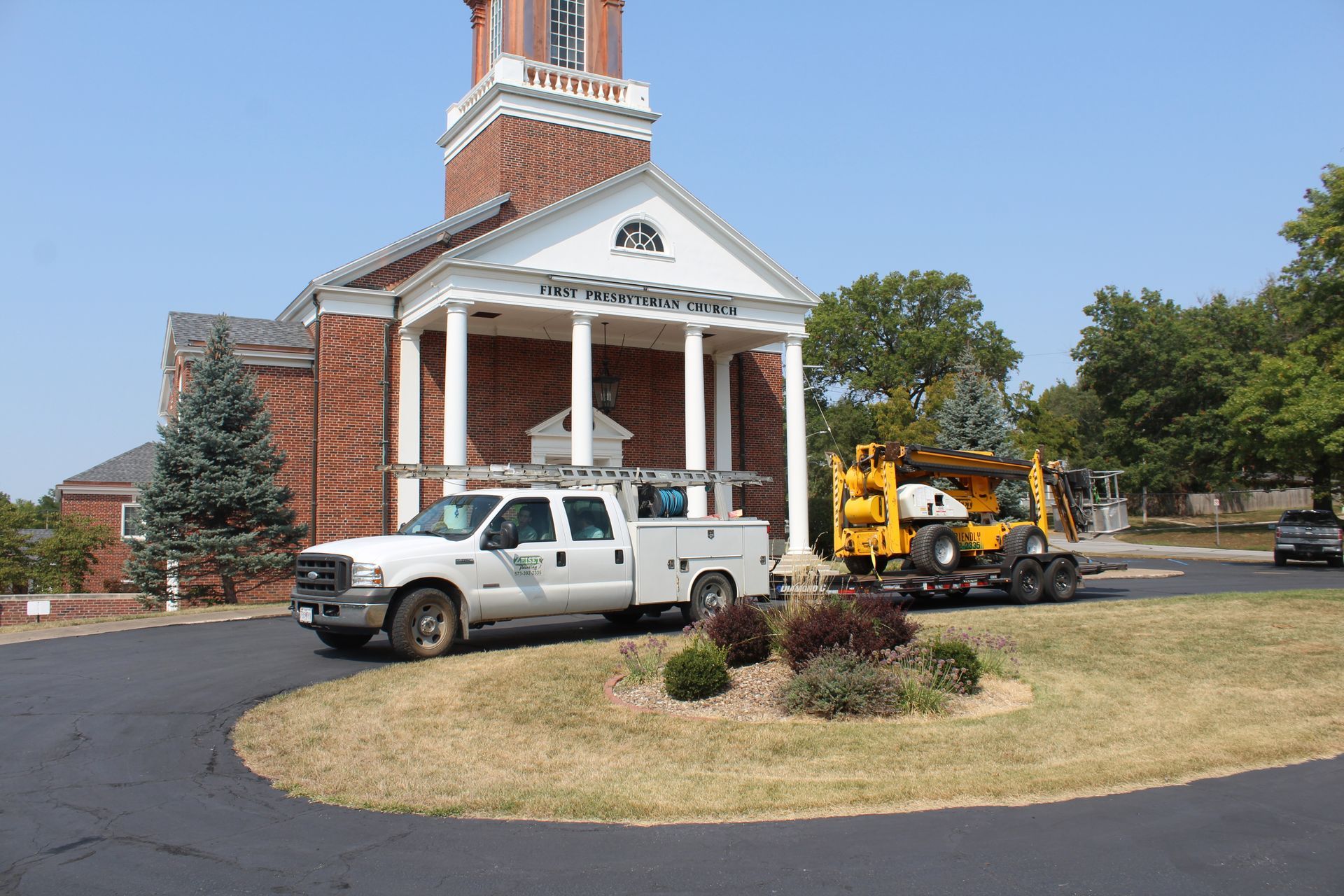 White pickup and yellow work truck parked by a brick building with white columns and a clock tower