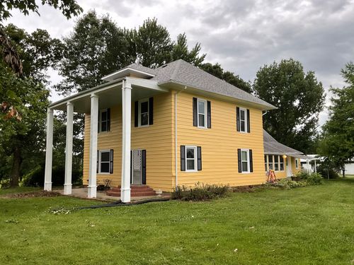 Yellow two-story house with white columns and shutters, standing in a grassy yard under cloudy sky