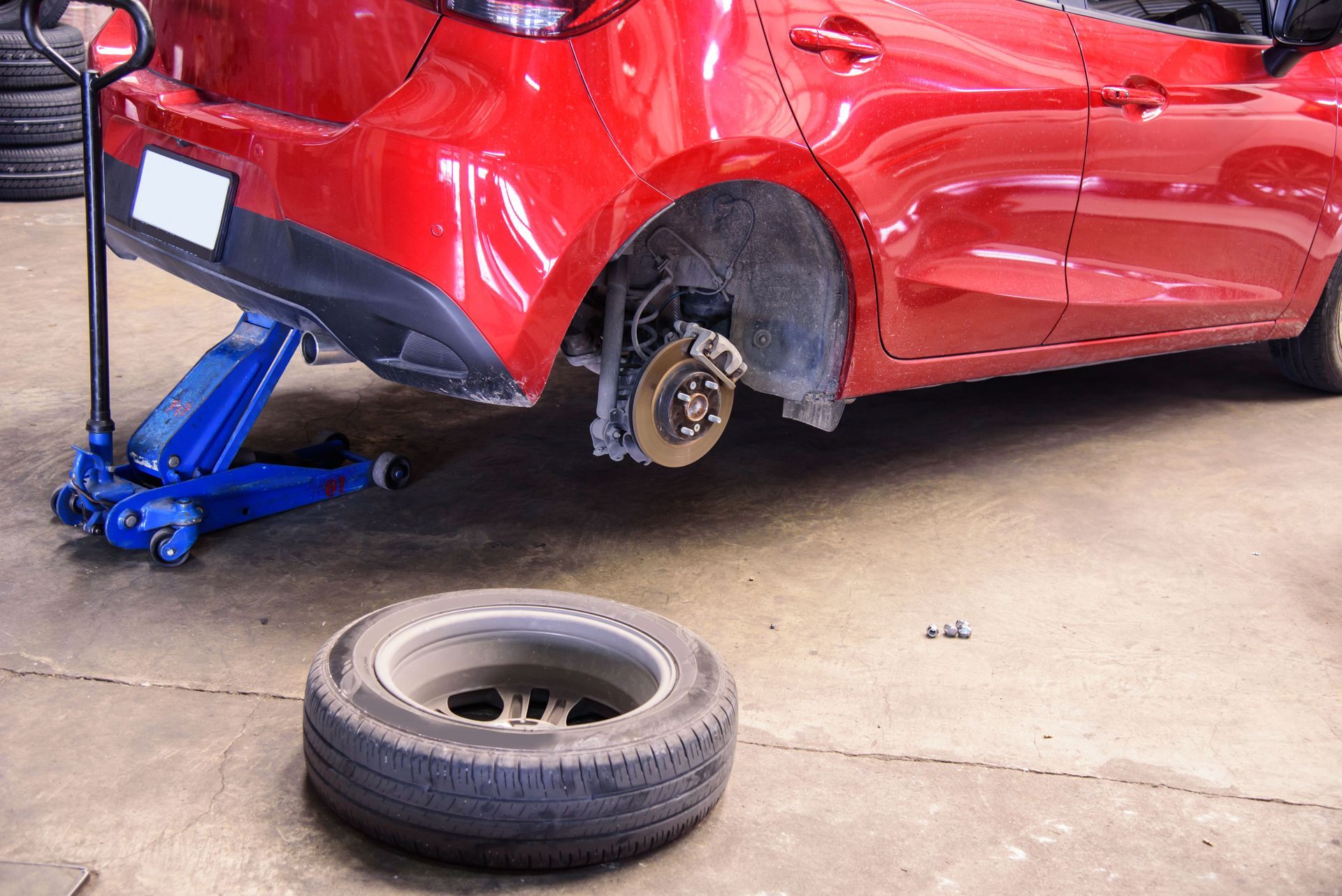 Red car lifted on a blue jack with a removed tire lying on the garage floor.