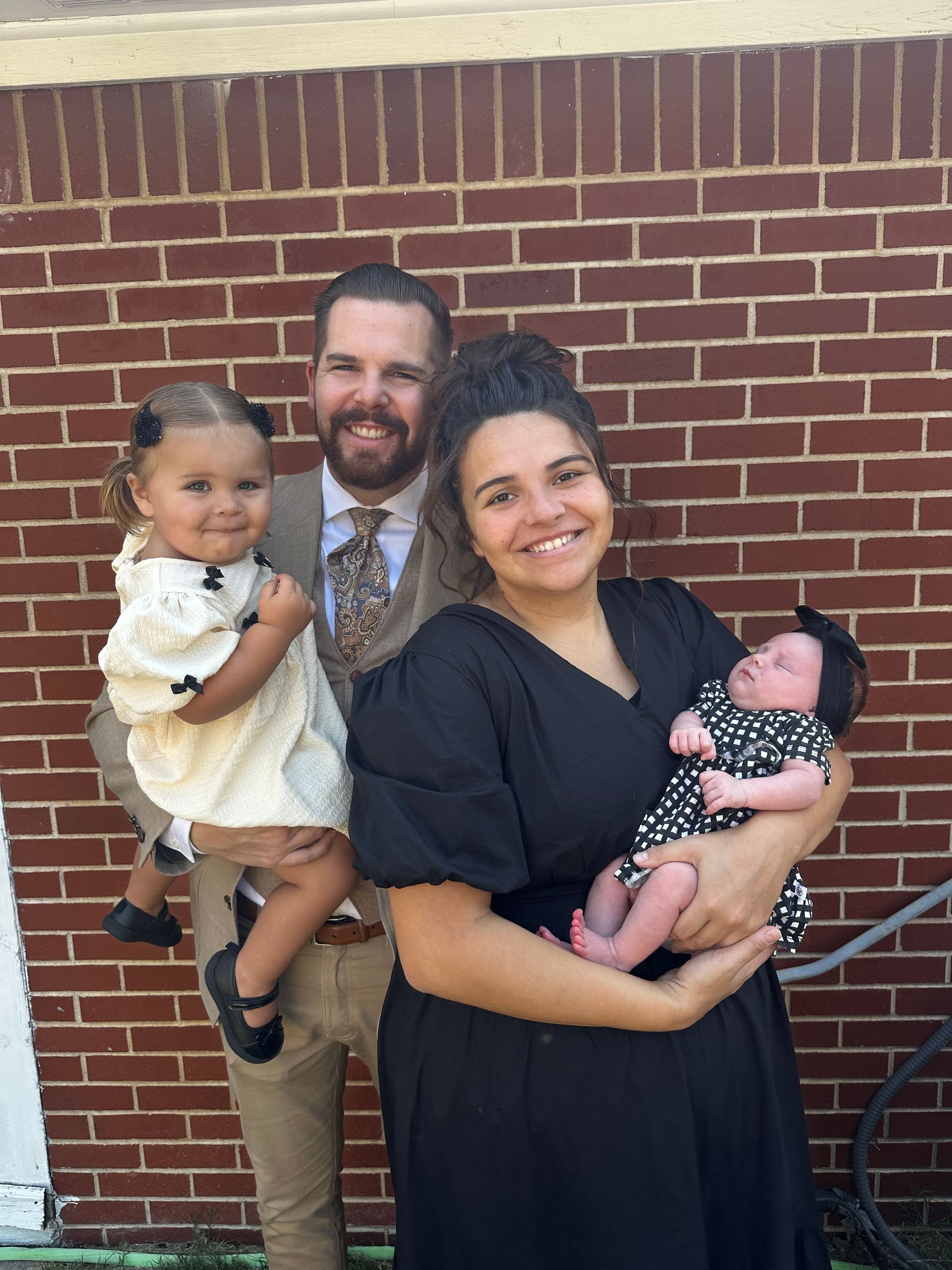 Family of four smiling outdoors in front of a brick wall, with two young children held in arms.