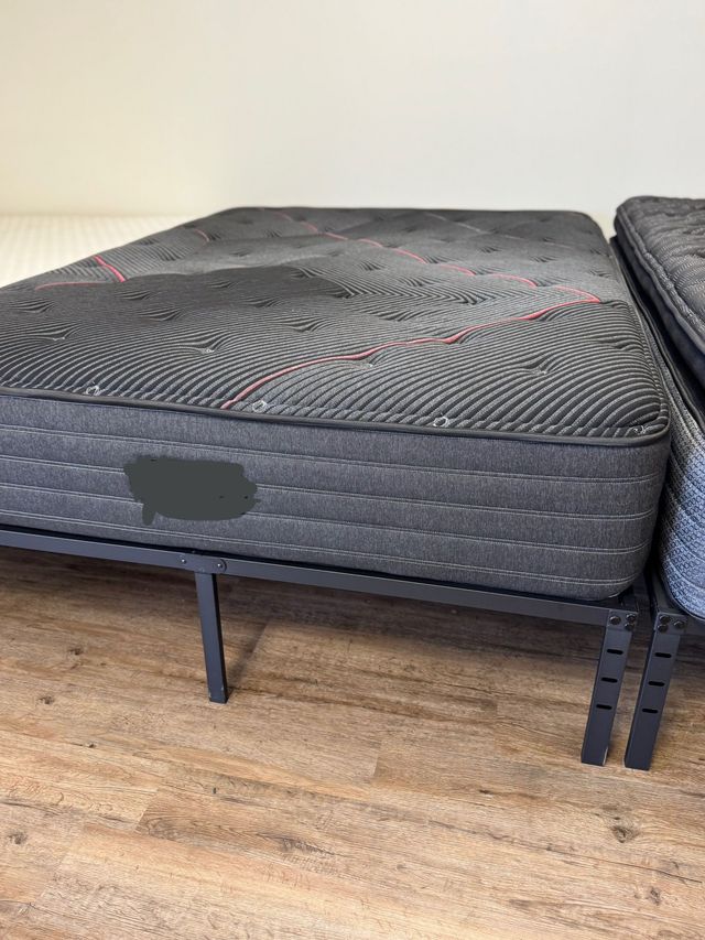 Black mattress on a metal bed frame in a sparsely furnished room with wooden floor.