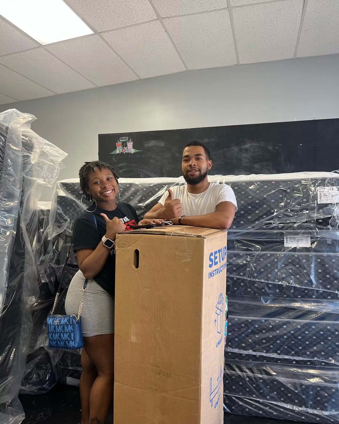Two people smiling beside a large cardboard box in a room with wrapped furniture.