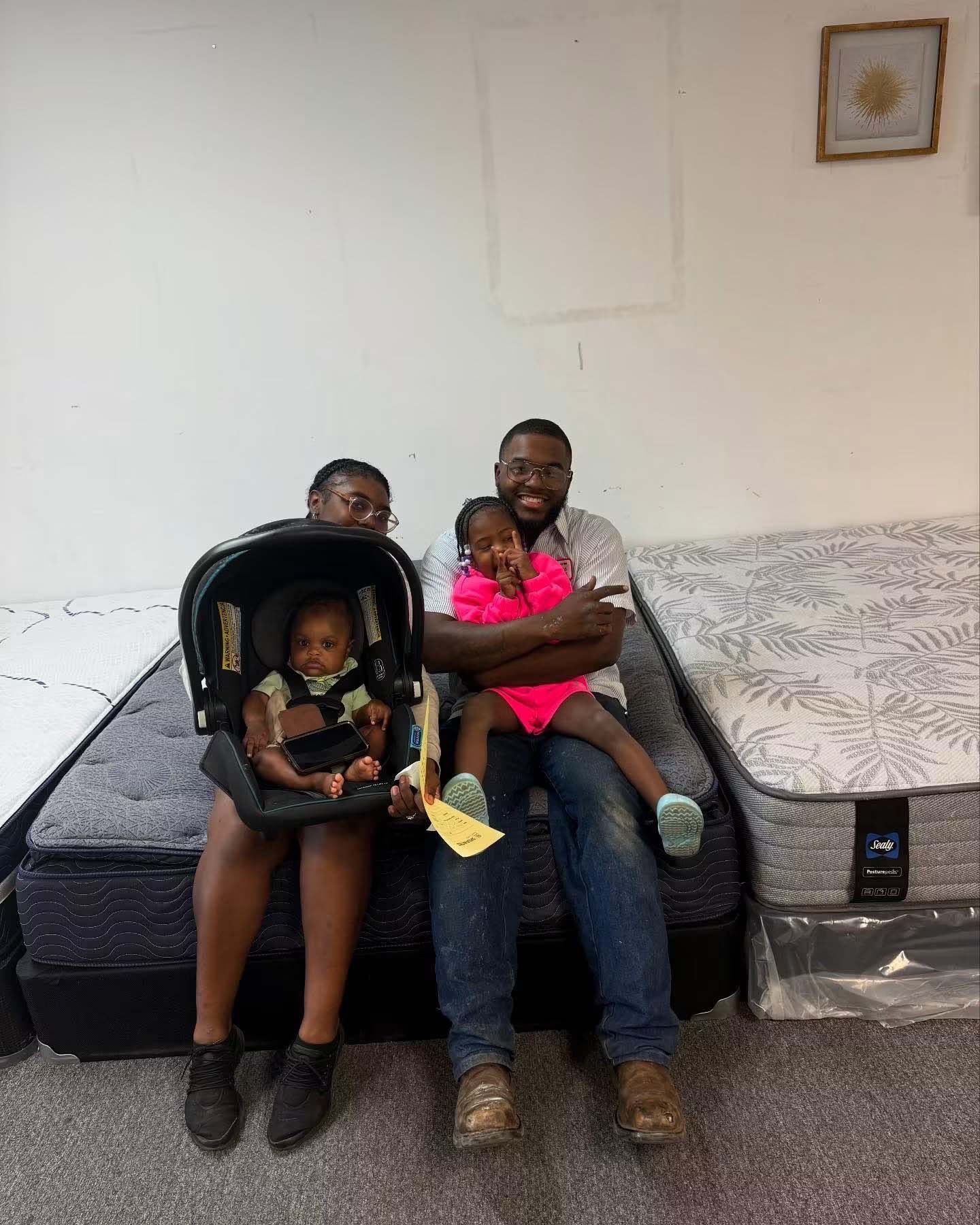 Family seated on a bed in a simple bedroom, two small children in car seats, one in a pink shirt and one on lap.