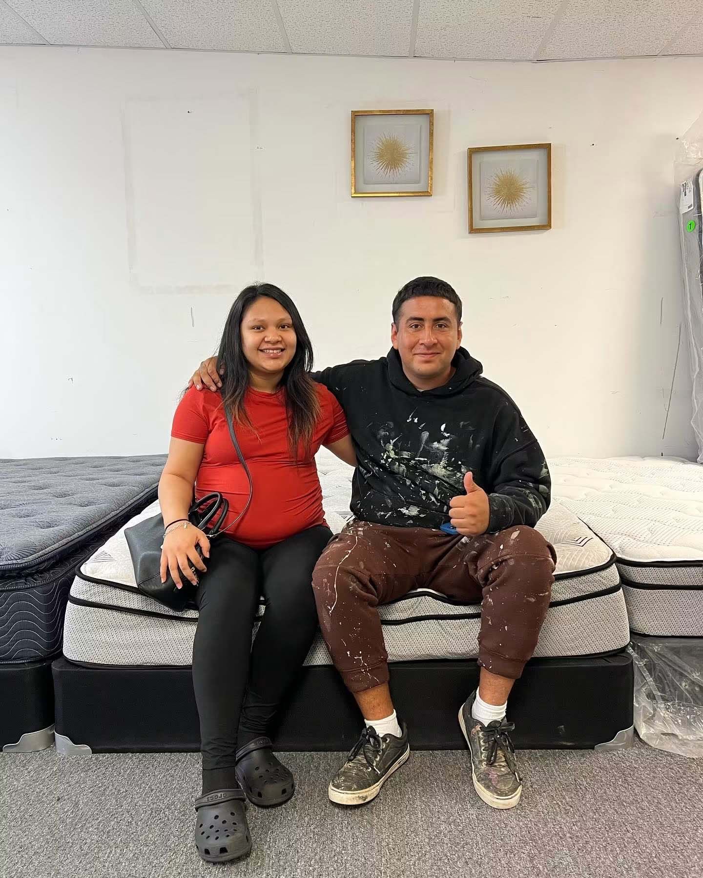 Two people sit on a bed smiling, with framed wall art behind them.