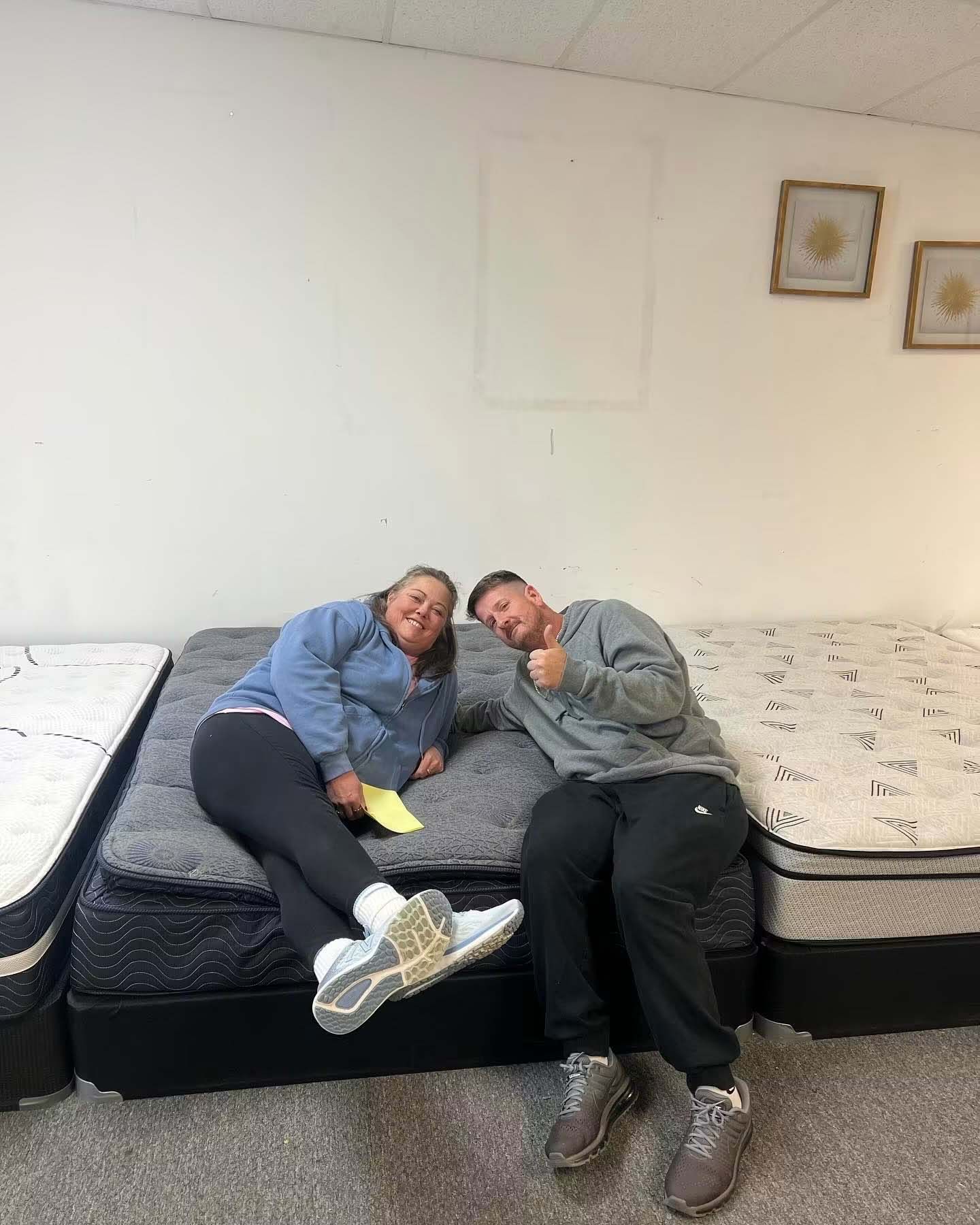 Two people sit close together on a bed in a mattress showroom, leaning in and smiling.