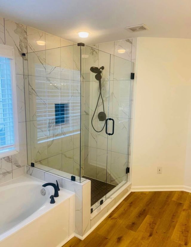 Bathroom with glass shower, white tub, and warm wood flooring in a bright corner room