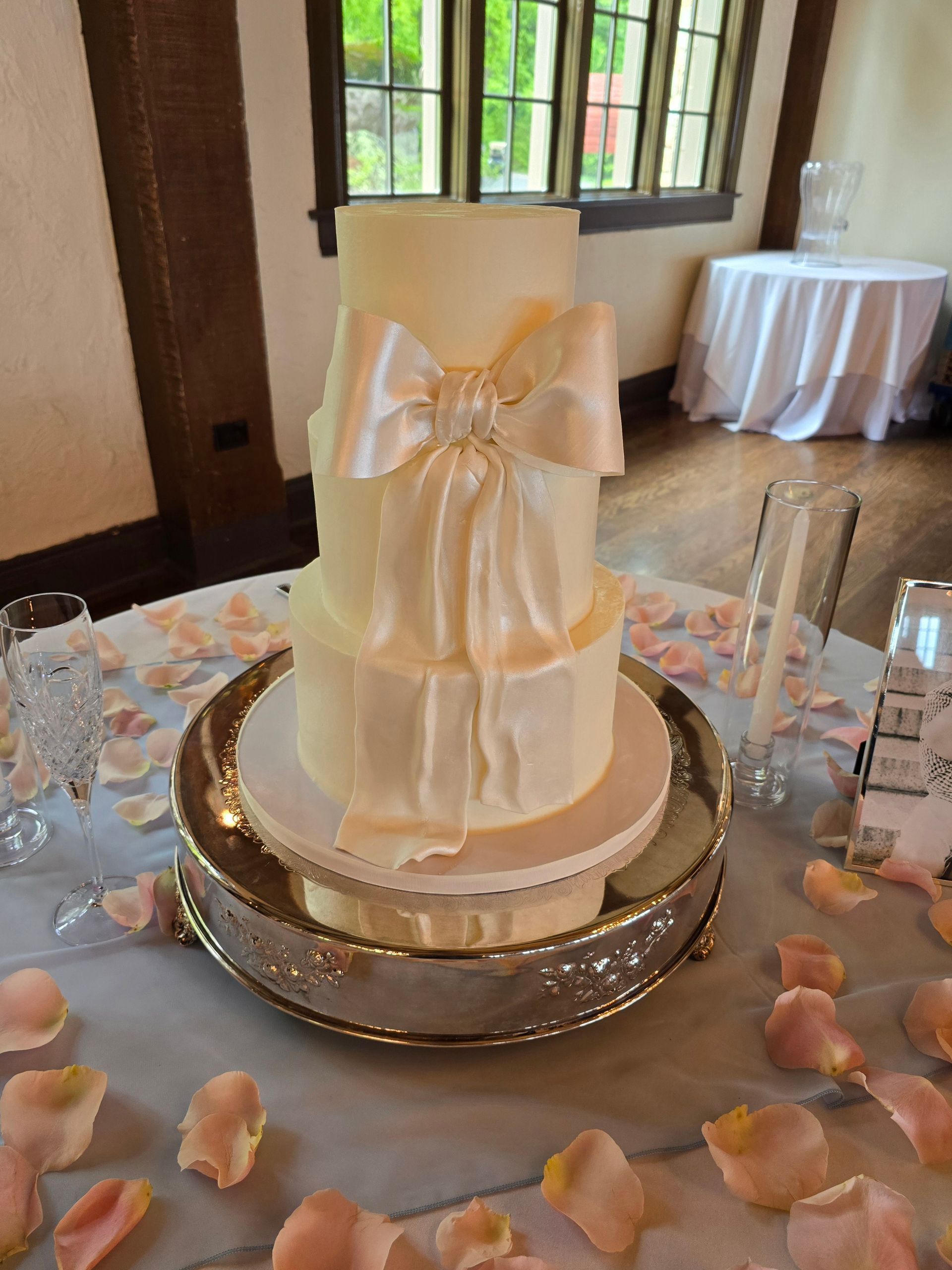 A white wedding cake with a bow is on a silver plate on a table.