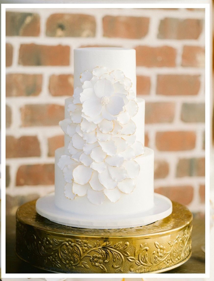 A white wedding cake is sitting on top of a gold cake stand in front of a brick wall.