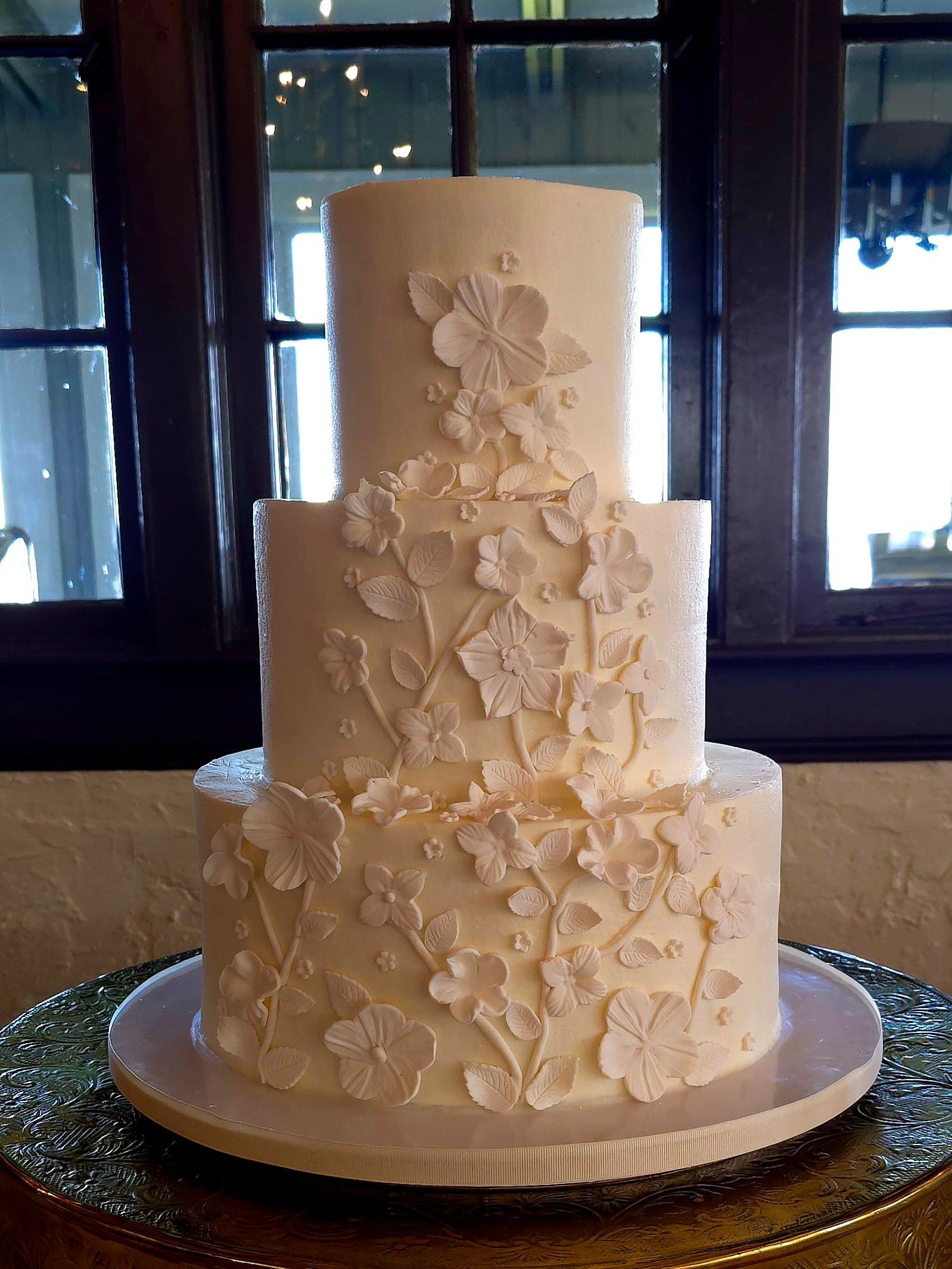 A white wedding cake with flowers on it is sitting on a table in front of a window.