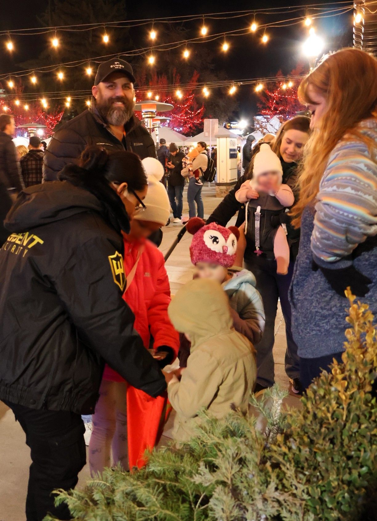 A group of people interact at a nighttime event illuminated by overhead string lights.