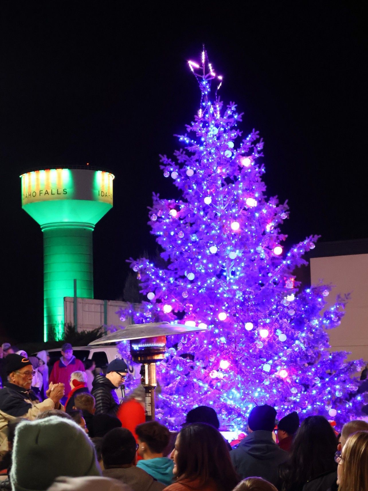 A tall, brightly lit purple Christmas tree stands outdoors at night next to a glowing green water tower.