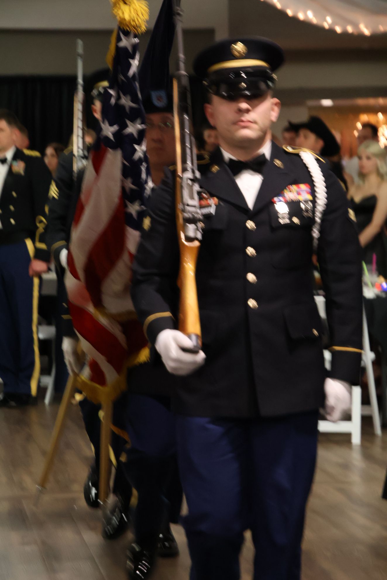 Members of a military color guard in dress blues carry the U.S. flag and a rifle at a formal event.