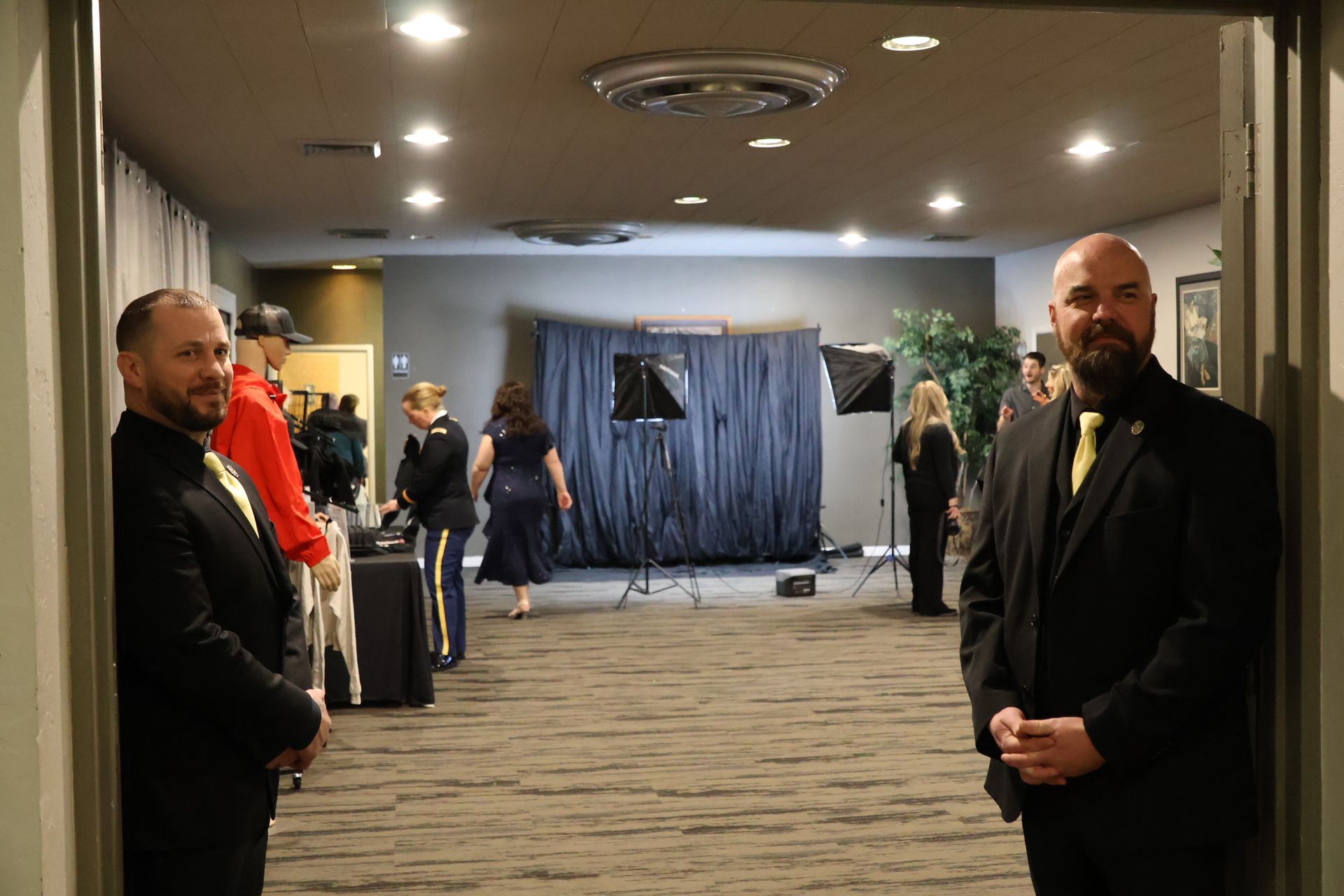 Two men in formal suits stand in a doorway facing a room with photography equipment and people setting up for an event.