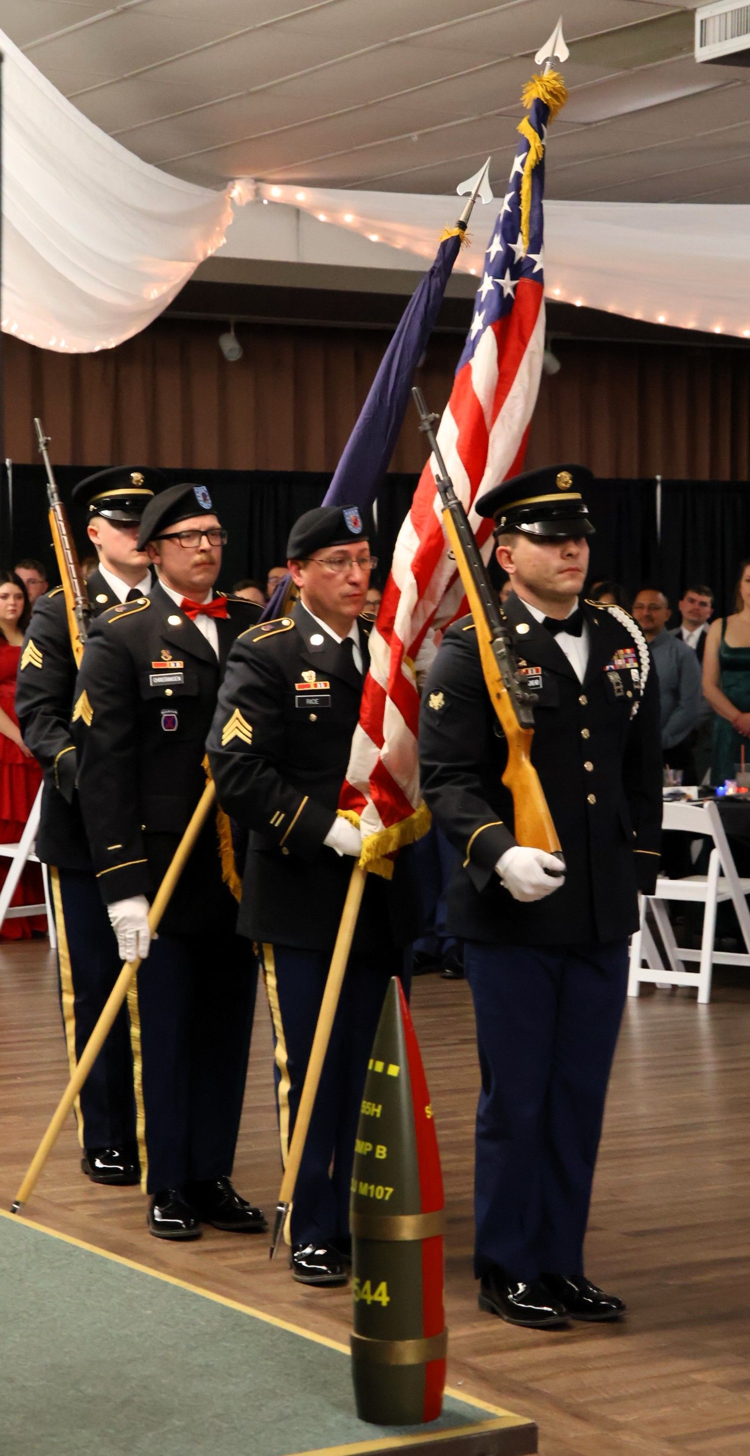 A color guard of four uniformed service members holds American and unit flags while standing at attention in a banquet hall.