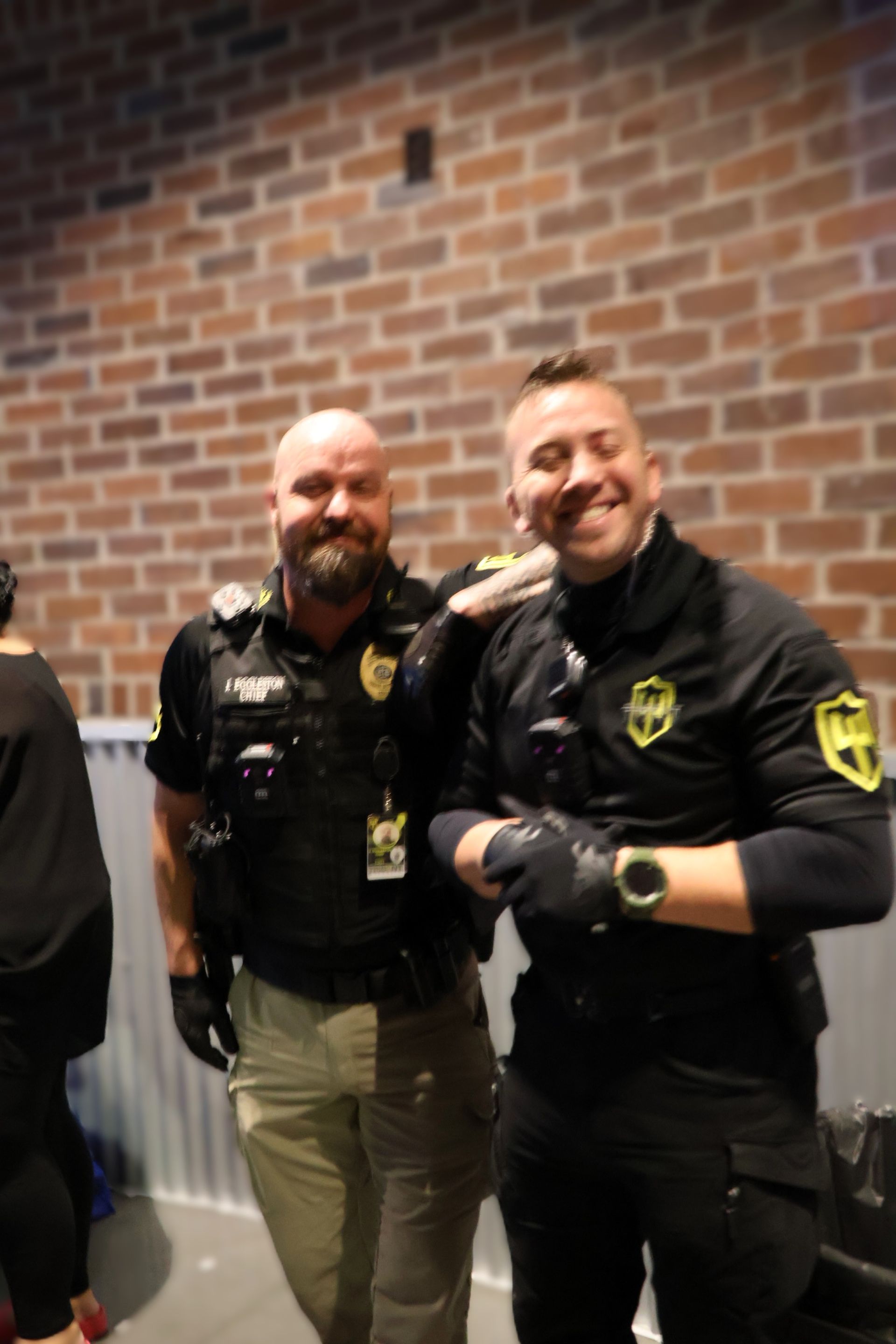 Two smiling security personnel in black uniforms and gloves pose together in front of a brick wall.