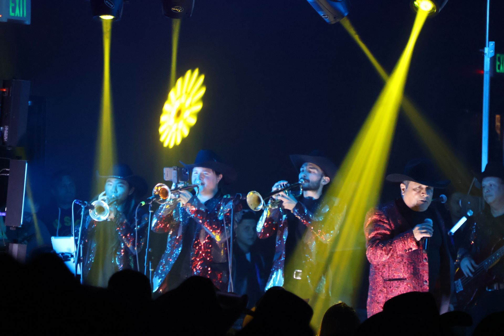 A band performing on stage in glittery outfits under yellow stage lights, with musicians playing brass instruments.