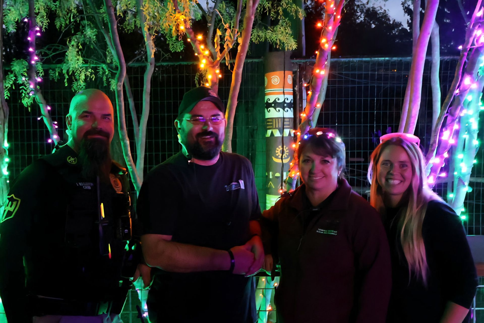 Four people stand smiling together outdoors at night, illuminated by colorful string lights hanging in the trees.