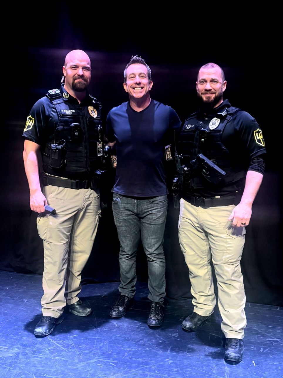 A smiling man stands between two uniformed law enforcement officers on a stage against a dark background.