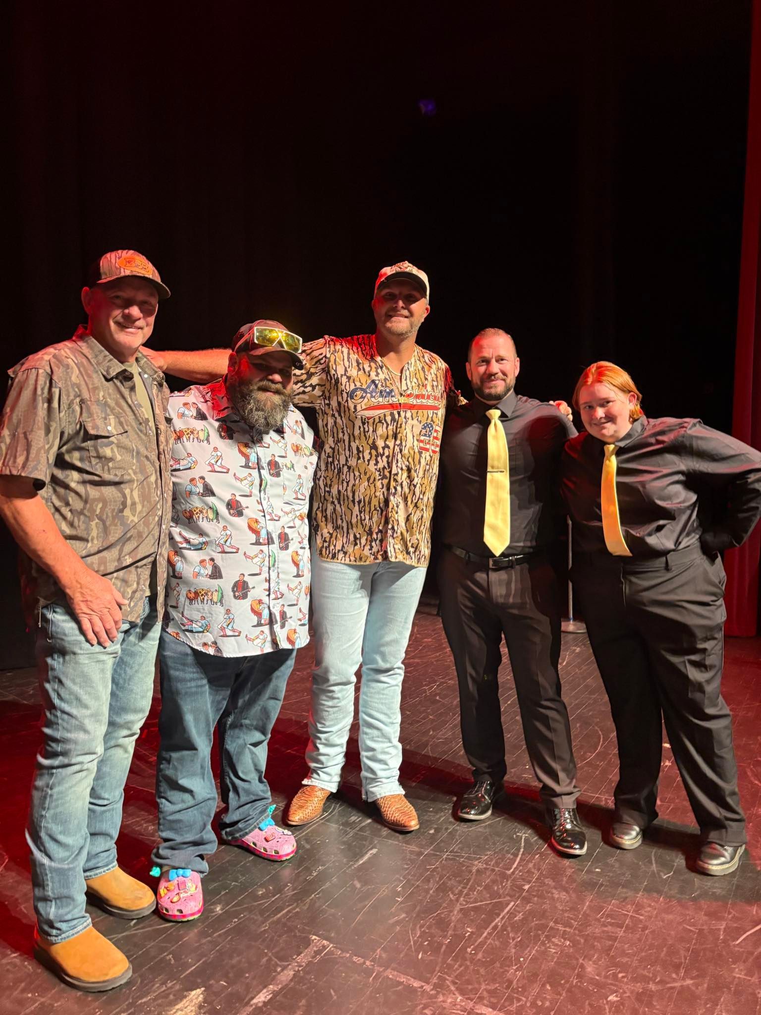 Five people stand in a line on a dimly lit theater stage, smiling and posing for a group photograph.